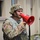 A U.S. Army soldier in full combat gear is speaking into a red megaphone/bullhorn, likely issuing commands or announcements. The soldier is wearing OCP (Operational Camouflage Pattern) uniform with a combat helmet and tactical vest. The setting appears to be a military installation or training facility.