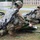 Two U.S. Army soldiers in full combat gear and ACU/OCP camouflage uniforms are performing tactical combat casualty care (TCCC) on simulated casualties lying on litters with emergency thermal blankets. The scene appears to be a training exercise conducted on a military installation, with military vehicles and buildings visible in the background. Both soldiers are armed and equipped with tactical gear while attending to the simulated wounded personnel.