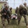 A group of U.S. Army soldiers in MultiCam/OCP camouflage uniforms are conducting a medical evacuation training exercise. Several soldiers are carrying a simulated casualty on a litter wrapped in a mylar/emergency blanket. At least one soldier is visibly identified as a MEDIC via a patch on their body armor.