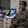 A U.S. Navy service member wearing digital camouflage NWU (Navy Working Uniform) and a tan/khaki face mask is seated in what appears to be an audiology or medical testing room. The individual displays rank insignia consistent with a Lieutenant (O-3) and a name tape partially visible on the uniform. Medical/audiological equipment including tablet-based diagnostic devices and a headset are visible on a cart beside them.