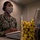 A U.S. Navy service member named Washington is seated at a workstation, wearing a camouflage uniform, a fabric face mask, and a headset. They appear to be working at a computer in what looks like an administrative or call center environment. A container filled with yellow foam earplugs is visible in the foreground.