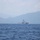 A white medium-to-large vessel, consistent in appearance with a coast guard or naval patrol cutter, is positioned on open blue water at mid-distance. A mountainous coastline shrouded in haze forms the backdrop. The image was taken from another vessel at sea level.