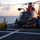 A U.S. Coast Guard helicopter, identifiable by its distinctive red and white color scheme, is on the flight deck of a Coast Guard cutter at sea during sunset or sunrise. A flight deck director in a yellow jersey and white helmet is signaling with a raised fist, while several crew members in blue uniforms work around the aircraft. The vessel is identified as 'VALIANT 621' based on text visible on the director's jersey.