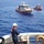 A U.S. Coast Guard operation is underway at sea, with personnel aboard a large white cutter vessel overseeing two smaller response boats in open blue water. A Coast Guard member in the foreground wearing a white hard hat, blue coveralls, and an orange life vest is managing a line connected to the boats ahead. The scene appears to depict a maritime training exercise or active rescue/law enforcement operation.