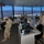 Military personnel in camouflage uniforms and face masks are stationed inside an air traffic control tower, overlooking a runway and airfield at dusk or dawn. The tower is equipped with multiple workstations, monitors, radios, and communication equipment. Several airmen appear to be conducting operations or receiving a briefing within the facility.
