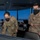 Two U.S. Air Force personnel wearing OCP (Operational Camouflage Pattern) uniforms and black face masks are working inside what appears to be an air traffic control tower. The individual on the right has a visible name tape reading 'GOLDSTEDT' and 'U.S. AIR FORCE' tape on their uniform. They are examining control equipment with a panoramic view of an airfield visible through the tower windows.