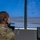 A military service member in camouflage uniform and face mask is stationed in what appears to be an air traffic control tower, monitoring the airfield. Multiple aircraft are visible on the tarmac through the large tower windows, with at least one aircraft airborne in the background. The controller has radar and communication equipment visible at the workstation.