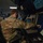 Two military personnel in OCP (Operational Camouflage Pattern) uniforms and black face masks are working at what appears to be an air traffic control or radar operations station. The foreground operator is pointing at a radar display screen showing aircraft tracks and navigational data. The environment is a dimly lit operations center with multiple monitors and control consoles.