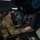 Military personnel in camouflage uniforms and face masks are working at a multi-screen control station in a dimly lit operations center. The foreground operator is seated at a console labeled 'ACTIVE BAY' with radar and data displays visible. Additional personnel are visible working at similar consoles in the background.