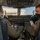 Two military aircrew members are seated in the cockpit of a large military transport aircraft, likely a C-17 Globemaster III, conducting pre-flight checks. Both individuals are wearing olive drab flight suits, headsets, and face masks. The right-seat crew member is holding what appears to be a checklist while the left-seat crew member interacts with cockpit controls.