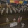 A military formation ceremony is taking place inside a large warehouse or hangar facility. Uniformed personnel wearing face masks are standing in organized rows facing officers at a podium area with an American flag, suggesting a formal military ceremony conducted during COVID-19 protocols. Several flags, including the American flag and what appear to be unit or organizational flags, are displayed in the background.