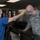 A healthcare worker in blue scrubs and a black face mask is using a non-contact infrared thermometer to take the temperature of a US Air Force personnel member wearing ABU (Airman Battle Uniform) camouflage and a face mask. The setting appears to be a medical facility or administrative building entrance, consistent with COVID-19 health screening protocols. A second military person in camouflage is visible in the background, also wearing a face mask.