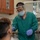 A dental professional wearing full PPE (face shield, surgical mask, glasses, green scrubs, and purple nitrile gloves) is performing a dental procedure on a seated patient. The setting is a clinical dental operatory with visible medical equipment and cabinetry. The practitioner is leaning toward the patient and holding a dental instrument near the patient's mouth.