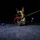 A Belgian Malinois or German Shepherd dog is photographed at ground level during nighttime, holding a red rope toy in its mouth. The dog is wearing a tactical harness and is attached to a bright yellow-green leash. The low-angle shot creates a dramatic perspective with distant lights visible in the dark background.