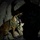 A military or law enforcement working dog, appearing to be a Belgian Malinois, is shown inside a dark cave or tunnel environment. The dog is equipped with a tactical vest/harness and leash, accompanied by a handler in camouflage clothing. The scene is illuminated by artificial light, suggesting a nighttime or underground operation.