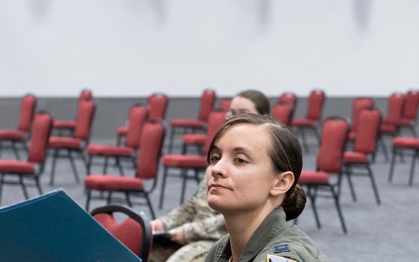 A female military aviator in an olive green flight suit sits in a conference or briefing room, extending a blue folder or binder toward someone off-camera. A second service member in camouflage uniform is visible seated in the background. The setting appears to be a large room with rows of red banquet-style chairs.