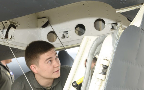 A young military personnel member in an olive/sage green flight suit is inspecting the underside of a military aircraft, examining what appears to be a landing gear bay or access panel area. At least one other person in similar attire is visible in the background. The setting appears to be an active airfield or flight line.