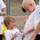 Two young children wearing white t-shirts interact outdoors during what appears to be a color run or powder paint event. The smaller child is pointing at and examining the color stains on the taller child's shirt, appearing excited and engaged. Both shirts show visible colored powder or paint marks, suggesting participation in a fun, messy activity.