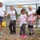 A group of children wearing white t-shirts are participating in an outdoor color/paint activity at what appears to be a school or daycare facility. The children are laughing and reacting with excitement as they are splattered with pink and blue paint or powder. A sign on the chain-link fence behind them reads 'School Age Care'.