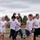 A group of children wearing white t-shirts are running enthusiastically outdoors in what appears to be an organized group activity or race. Several adults are visible in the background, seemingly supervising or participating. The setting is an open, semi-arid landscape with scattered pine trees under an overcast sky.