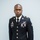 A male soldier in U.S. Army dress blue uniform stands in a formal portrait pose against a light gray background. The uniform displays multiple insignia, badges, ribbons, and a name tape reading 'LOUIGENE'. The image appears to be an official or semi-official military portrait photograph.