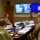 A group of U.S. Army soldiers in camouflage uniforms are seated around a conference table participating in a video teleconference meeting. A soldier appearing on a wall-mounted screen is addressing the group remotely, while a second monitor displays a Windows 10 desktop with what appears to be a U.S. Army seal. The setting is a formal military conference room with binders, documents, and name placards on the table.