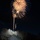 A nighttime photograph captures fireworks exploding above Mount Rushmore National Memorial in South Dakota. The iconic granite sculpture featuring four U.S. presidents is illuminated from below against a dark sky. The long-exposure shot captures multiple firework bursts in gold, white, and blue tones rising from behind the monument.