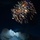 A nighttime photograph captures Mount Rushmore National Memorial illuminated by artificial lighting, with a spectacular fireworks display bursting in the sky above. The four presidential faces carved into the granite mountain are clearly visible against the dark blue night sky. This appears to be a Fourth of July or patriotic celebration event.