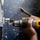 A person is using a yellow and black DeWalt cordless drill to work on a metal bolt or threaded fastener mounted on a painted metal surface. The work surface appears to be a dark workbench or floor with paint splatters. The scene suggests mechanical repair or assembly work in an industrial or workshop environment.