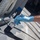 A person wearing a blue nitrile glove is applying dark gray or black paint to a gray metal surface using a paintbrush. The setting appears to be on or near a vessel or maritime structure, evidenced by the thick white rope and gray painted metal surfaces visible in the frame. Blue painter's masking tape is visible in the background, indicating deliberate, careful paint work.