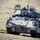 An armored fighting vehicle, consistent with an M2 Bradley Infantry Fighting Vehicle, is traversing open grassland terrain with its headlights illuminated. A crew member is visible in the open commander's hatch wearing a helmet and protective eyewear. The vehicle appears to be equipped with additional armor packages and electronic systems mounted on the turret.