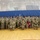 Korea's Top Dentist Changes Command A large group of U.S. Army soldiers in camouflage uniforms and black berets pose for a formal unit photograph inside a gymnasium or indoor sports facility. Three soldiers are kneeling in the front row, with one holding a maroon unit guidon flag. Flags are visible in the background, suggesting this may be a unit activation, change of command, or similar military ceremony.
