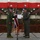 Two military personnel in green flight suits are posing together while jointly holding a certificate or award document inside a large aircraft hangar. Between them stands a United Kingdom flag on a stand, with a United States flag visible in the background, suggesting a joint US-UK military ceremony. The setting appears to be a formal military recognition or award presentation event.