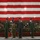 Four military personnel stand at attention on a raised platform beneath a large American flag displayed in what appears to be a military hangar or large indoor facility. Three individuals wear olive green flight suits with military patches, while one person in the center-rear wears a camouflage uniform and holds ceremonial flags. The setting suggests a formal military ceremony, likely a change of command or unit activation event.