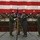 A military change of command ceremony is taking place inside a large aircraft hangar decorated with an oversized American flag backdrop. Two officers in green flight suits are exchanging a unit guidon (flag on a staff), which is the central ritual of a change of command ceremony. Additional personnel in both flight suits and camouflage uniforms are present as witnesses.