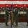 A military change of command ceremony is taking place inside a large hangar decorated with an American flag backdrop and camouflage netting. Two officers in green flight suits are exchanging a unit guidon (flag), which is the central symbolic act of a change of command ceremony. A third officer stands at attention to the right, and a soldier in camouflage uniform holds additional flags in the background.