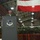 A U.S. Air Force officer in flight suit and service cap stands at a podium delivering a speech inside what appears to be an aircraft hangar. The podium bears a shield emblem with the Statue of Liberty and the text 'STATUE DE LA LIBERTÉ,' suggesting a unit or wing insignia. A large American flag is draped in the background above camouflage netting.