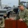 EODESU One Holds Change of Command A U.S. Navy officer in digital camouflage (NWU Type III) uniform is speaking at a wooden podium with a microphone. The name tape on his uniform reads 'ROJAS' and he displays a Captain's insignia (eagle device). A second uniformed Navy personnel wearing a black face mask is seated in the background.