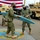EODESU One Holds Change of Command Two U.S. Navy personnel in camouflage uniforms and face masks are handling a turquoise surfboard during what appears to be a military ceremony. They are standing on a U.S. Navy Explosive Ordnance Disposal Expeditionary Support Unit seal/emblem on the ground. The setting includes military vehicles (MRAPs/HMMWVs), a large American flag in the background, and one service member is wearing a military medal.