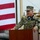 EODESU One Holds Change of Command A U.S. Navy officer named Pavlik is speaking at a podium during what appears to be an official military ceremony. He is wearing Navy Working Uniform (NWU) camouflage with a medal visible on his chest, standing before a large American flag. The setting appears to be an outdoor naval or military installation.