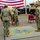 EODESU One Holds Change of Command A U.S. Navy military ceremony is taking place outdoors, likely a change of command or similar formal event for the Explosive Ordnance Disposal Expeditionary Support Unit. Three Navy personnel in camouflage uniforms are present, with one rendering a salute. The scene is set against a large American flag backdrop with military vehicles in the background.