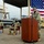 EODESU One Holds Change of Command A U.S. Navy military ceremony is taking place outdoors at what appears to be a naval installation. One uniformed sailor stands at a wooden podium speaking into a microphone, while two other sailors are seated in red chairs behind him. A large American flag serves as a backdrop, and military equipment including rigid inflatable boats and a Humvee are visible in the background.