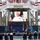 A military ceremony is taking place at the National Naval Aviation Museum, with a small socially-distanced audience seated in chairs facing a stage. A large LED screen displays a Navy officer in white dress uniform delivering remarks remotely via video, with a MyNavyHR logo visible. The venue is decorated with red, white, and blue bunting and features multiple military flags on stage.