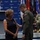 A military ceremony appears to be taking place in a formal venue with flags and a podium in the background. A woman in a black dress, wearing a medal on her chest, is interacting with a military officer in an olive green flight suit bearing a US flag patch. A senior military officer in camouflage uniform is seated on stage in the background.