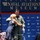 A military aviator in an olive green flight suit is being embraced by a woman in a navy blue dress at what appears to be a formal ceremony at the National Naval Aviation Museum. The man is using a walker (branded 'Drive'), suggesting he is recovering from an injury or medical condition. The emotional moment appears to take place on a stage with military branch insignia visible on the backdrop.