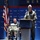 A military officer in an olive green flight suit and white garrison cap is speaking at a podium during a formal event at the National Naval Aviation Museum. A second individual in a similar flight suit is seated in a chair with a walker nearby, suggesting a medical condition or injury. The backdrop features the museum's branded step-and-repeat banner along with an American flag.