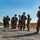 A group of military personnel in digital camouflage uniforms are standing on a gravel surface in an outdoor field environment. They appear to be in a formation or waiting posture, with several carrying rifles. The scene suggests a training exercise or field operation, likely at a military base or range.