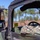A military service member in MARPAT camouflage uniform is seated in the driver's seat of a military tactical vehicle, likely a HMMWV (Humvee). A second service member is visible in the side-view mirror reflection, also wearing MARPAT camouflage and a cover. The scene appears to be an outdoor training environment with red dirt terrain and pine trees in the background.