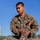 A U.S. military service member wearing MARPAT camouflage uniform with the name tape 'MAMASIG' is performing what appears to be a combat casualty care or first aid drill on another service member lying on the ground. The standing individual is kneeling over the casualty and appears to be assessing or treating the chest/torso area. This appears to be a training exercise conducted outdoors under clear blue skies.