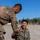 Two U.S. Marines in MARPAT digital camouflage uniforms are interacting outdoors on a sandy training area. One Marine, identified by the name tape 'BARBOSA' and 'U.S. MARINES' tape, is seated on the ground smiling broadly while another Marine leans down extending a hand toward him. The setting appears to be a field training exercise in a dry, open environment with trees visible in the background.